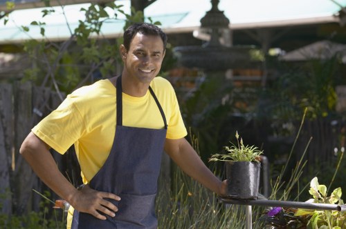 A man wearing a yellow t-shirt and a navy gardening apron is standing in a well-maintained garden in an outdoor space, surrounded by greenery and plants. He is smiling at the camera and holding a black plastic pot containing a small plant, which he appears to be preparing for planting or watering. The garden features a lush lawn with dense grass, bordered by a variety of shrubs and flowering plants, including some purple and white blossoms. In the background, there are trees and garden structures, such as a gazebo or pergola, partially visible through the leafy environment. The surfaces include a paved area or stone patio nearby, providing a clean and organized outdoor environment suitable for gardening and landscaping activities. The weather appears clear with natural daylight illuminating the scene, suitable for outdoor gardening work. This image relates to professional gardening services in Aldgate, highlighting garden maintenance, planting, and outdoor care provided by Garden Maintenance Aldgate, a local company specializing in comprehensive lawn, shrub, and flower bed maintenance.