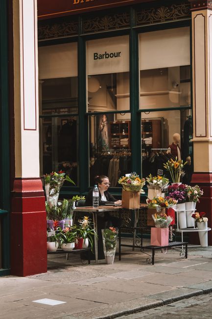 A flower vendor's outdoor stall located on a city sidewalk in front of a shop window displaying the 'Barbour' logo. The stall features a variety of colorful flowers and plants arranged in white, pink,