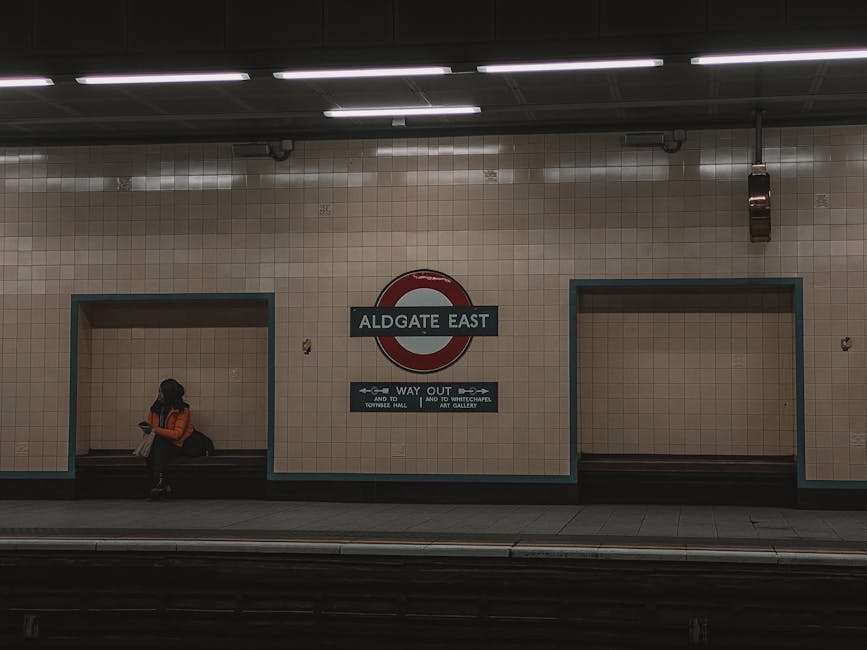 A woman sitting on a bench at Aldgate East underground station platform, with beige tiled walls and a large round station sign reading 'ALDGATE EAST' in the center. She is wearing an orange jacket and