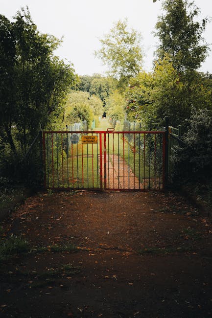 A red metal gate with vertical bars blocks the entrance to a lush, green garden path, indicating restricted access for maintenance work. The ground in front of the gate is dark, damp soil, transitioni