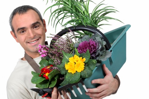 A man with a friendly smile is holding a light blue gardening trowel and a matching rake, each filled with various colorful plants and flowers, in an outdoor garden setting. Behind him, there is a lush green potted plant with long, narrow leaves, and the background features a well-maintained grassy lawn with a mixture of dense green grass and soil patches. The garden appears to be part of a landscaped backyard or front yard, with a clean, paved pathway visible in the distance. The scene is brightly lit with natural sunlight, indicating clear weather. The image reflects professional gardening and landscaping activities, emphasizing plant care, lawn maintenance, and outdoor beautification, consistent with services offered by companies like Garden Maintenance Aldgate in the London area, close to the postcode EC3 or EC4.