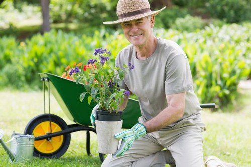 A mature man wearing a straw hat, light grey t-shirt, and gardening gloves is kneeling on a well-maintained lawn, holding a potted plant with flowering purple and red blooms. Behind him, a bright green wheelbarrow with yellow wheels is partially visible, loaded with gardening tools and additional potted plants, set on a lush, green outdoor space. The garden features a grassy lawn with tidy edges, adjacent to a dense hedge and various shrubs, indicating a neatly landscaped garden environment. In the background, tall trees and natural foliage create a shaded, tranquil atmosphere, with dappled sunlight filtering through the leaves. This scene exemplifies careful outdoor maintenance and planting activities typical of professional gardening services in Aldgate, connecting to the broader context of lawn and garden care, landscaping, and planting services available locally. The overall scene suggests a well-kept, healthy garden during daylight, with vibrant natural tones and a quiet, orderly outdoor setting reflecting the expertise of Garden Maintenance Aldgate in maintaining thriving gardens in the area.