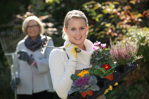 A young woman with blonde hair, wearing a white sweater and yellow gardening gloves, is standing outdoors in a garden during daylight, holding a black tray filled with colorful flowering plants, including purple, pink, and red blooms, with green foliage. In the background, an older woman with glasses, a white coat, and a scarf is smiling and holding a garden rake, standing amidst a lush garden with various shrubs, trees, and flower beds. The garden features well-maintained grassy areas, soil patches, and paved pathways, with sunlight filtering through the trees, creating a warm and inviting outdoor environment suitable for professional gardening and landscaping care by Garden Maintenance Aldgate, focused on enhancing outdoor spaces in the local area.